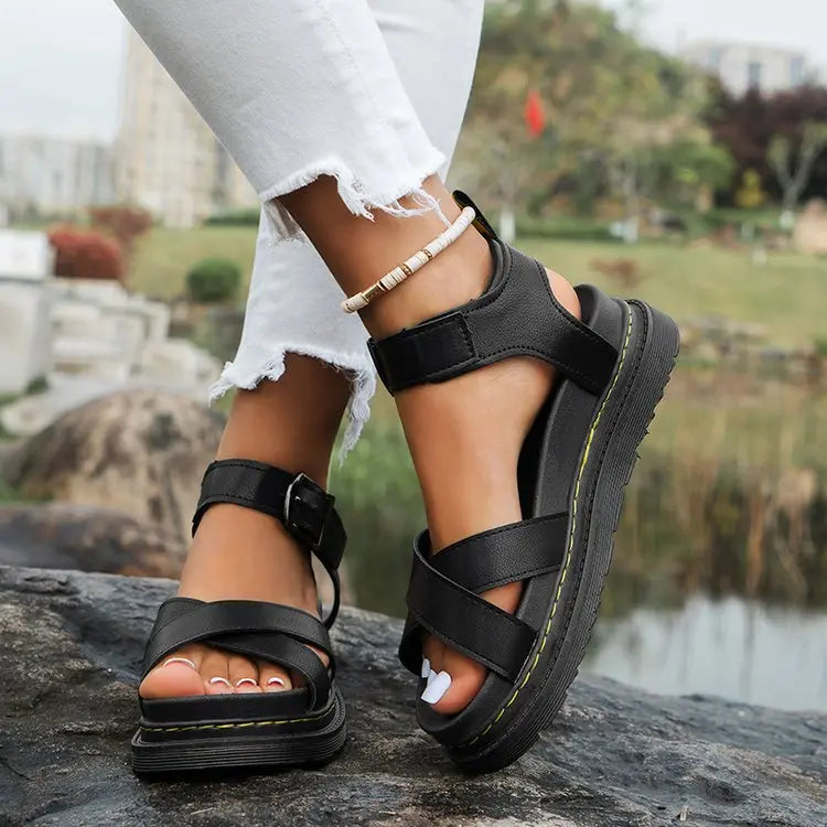 Black chunky platform sandals featuring criss-cross straps and an adjustable ankle buckle, shown on a model's feet styled with ripped white jeans in an outdoor, natural setting.