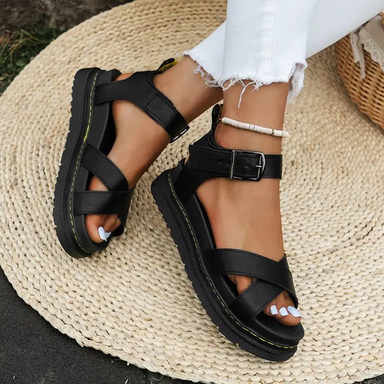 Black chunky platform sandals with criss-cross straps and buckle ankle closure, worn by a model with ripped white jeans on a woven mat.