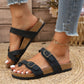 Woman's feet wearing black suede-effect cork footbed slide sandals with adjustable antique brass buckles, paired with ripped jeans and an anklet.