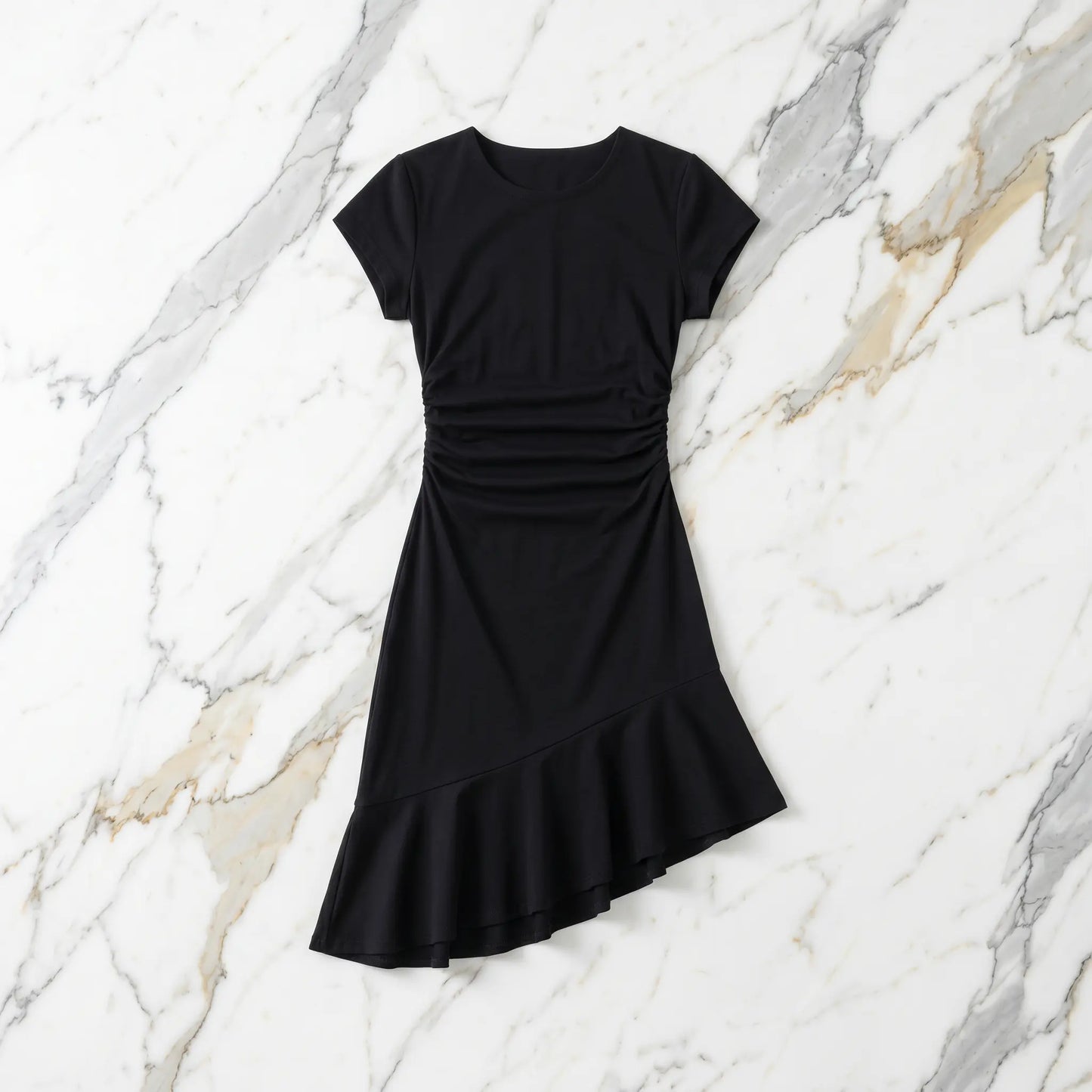 Black premium cotton cap-sleeve dress featuring a crew neck, right-side waist ruching, and an elegant asymmetrical ruffled hem. Laid flat on a white marble background.