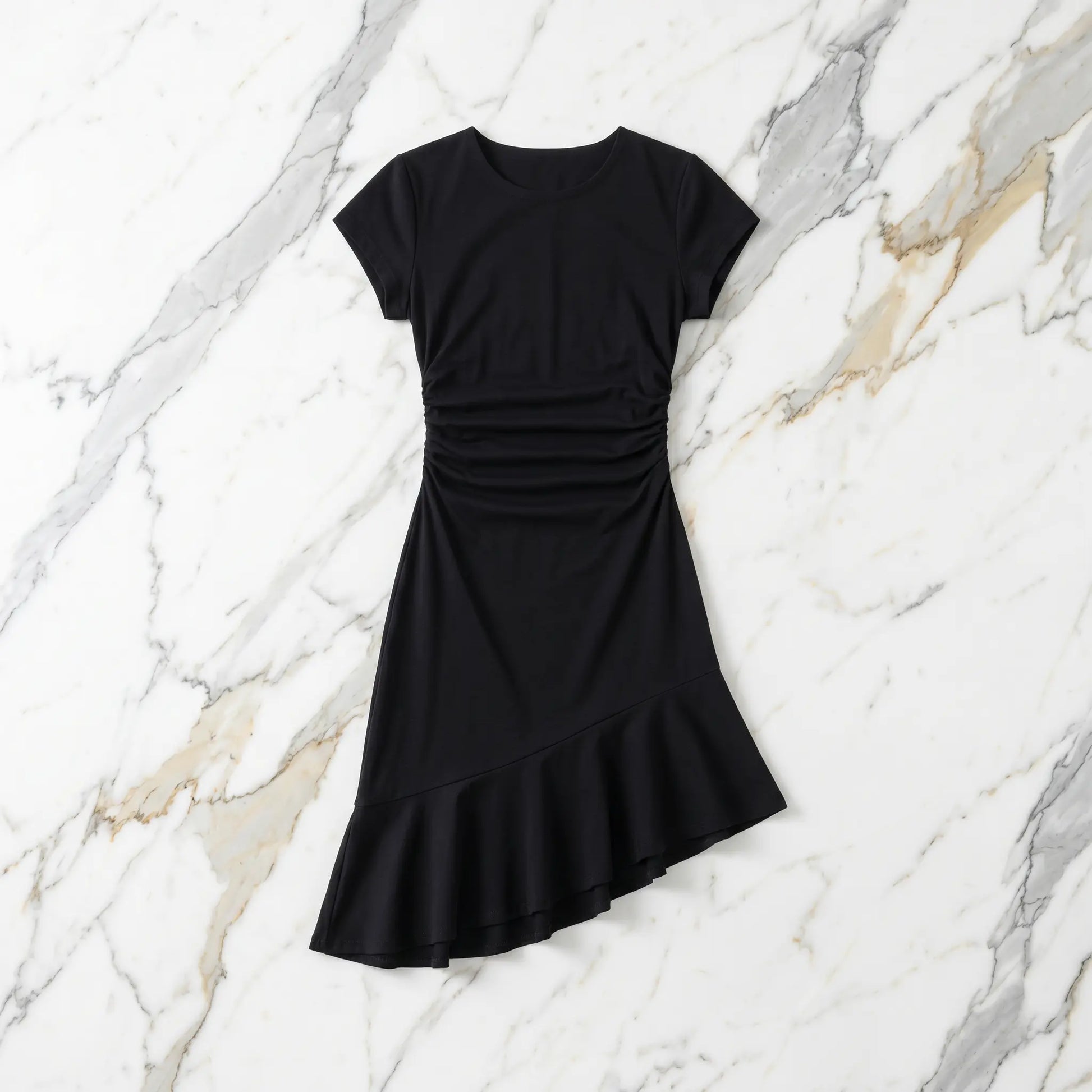 Black premium cotton cap-sleeve dress featuring a crew neck, right-side waist ruching, and an elegant asymmetrical ruffled hem. Laid flat on a white marble background.