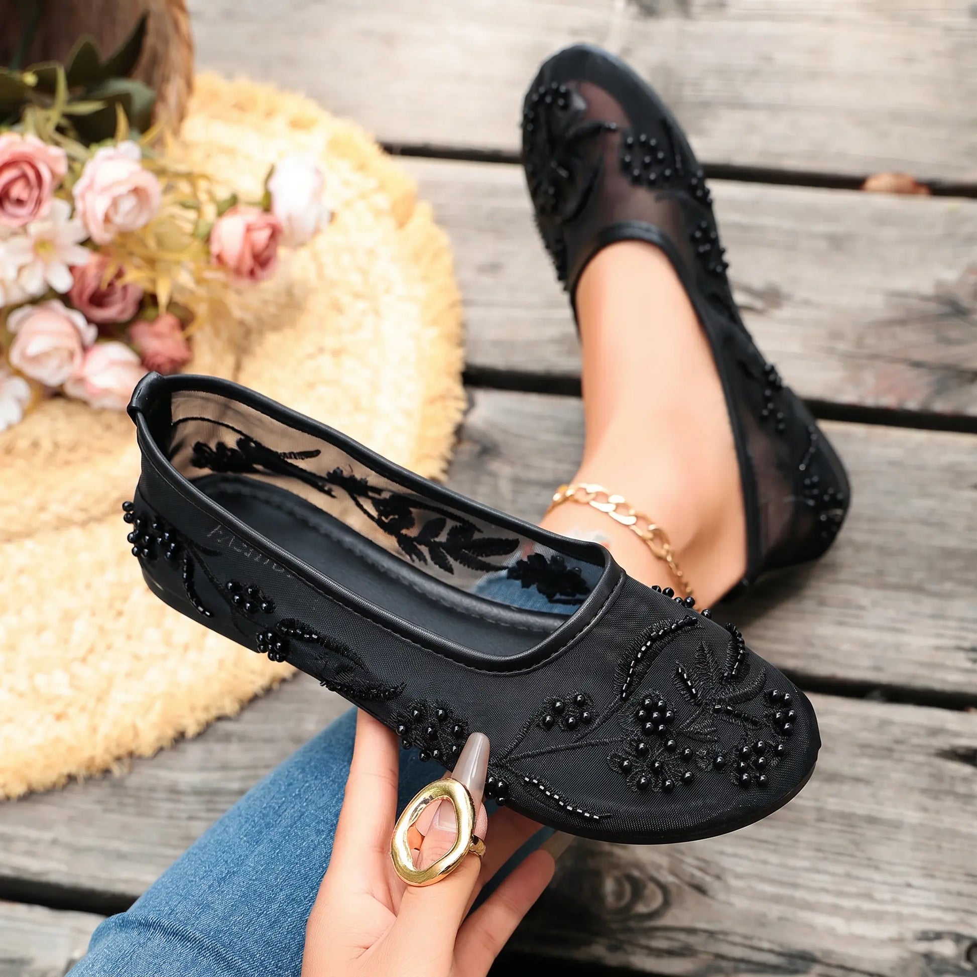 Black sheer mesh ballet flats embellished with delicate floral embroidery and faux pearls, styled on a foot with a gold anklet and held in hand on a wooden plank.