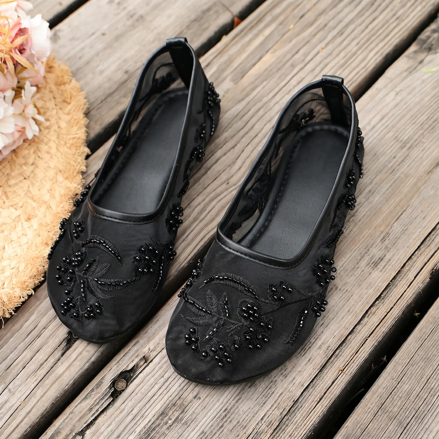 Black Lyraen embellished mesh ballet flats with intricate floral embroidery and pearl details, shown from a three-quarter angle on a weathered wooden surface with a straw hat.