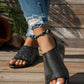Woman's feet wearing black embellished peep-toe flat sandals with a single strap, styled with ripped blue jeans and a turquoise beaded anklet on a wooden deck.