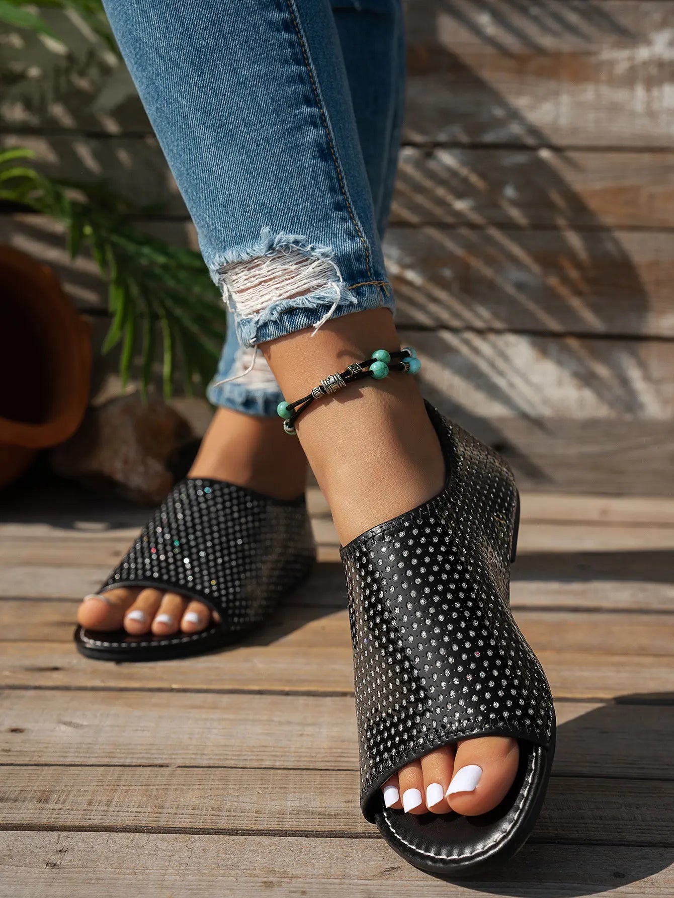 Woman's feet wearing black embellished peep-toe flat sandals with a single strap, styled with ripped blue jeans and a turquoise beaded anklet on a wooden deck.