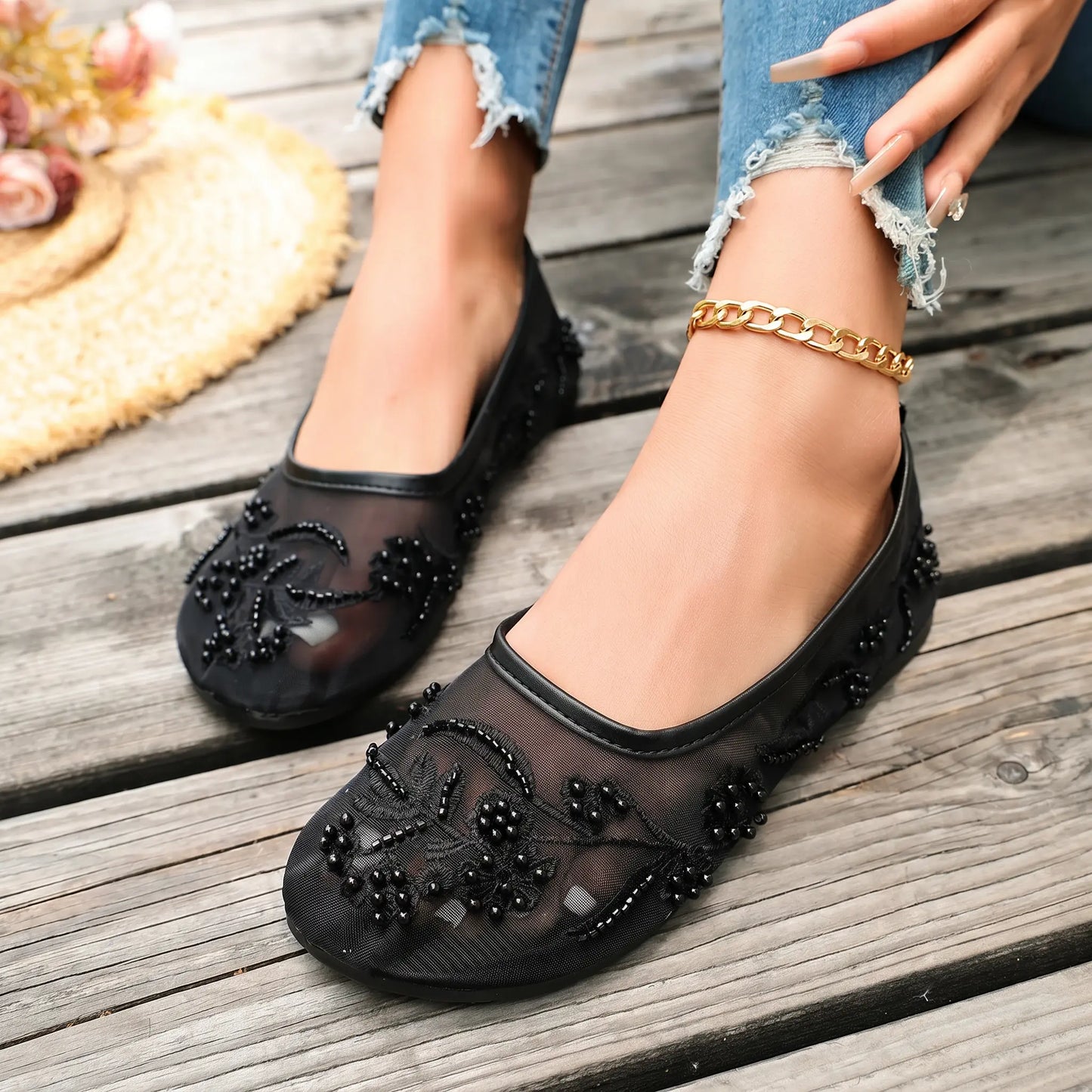 Black sheer mesh ballet flats embellished with intricate pearl and bead floral embroidery, worn by a model with distressed jeans on a wooden plank surface.