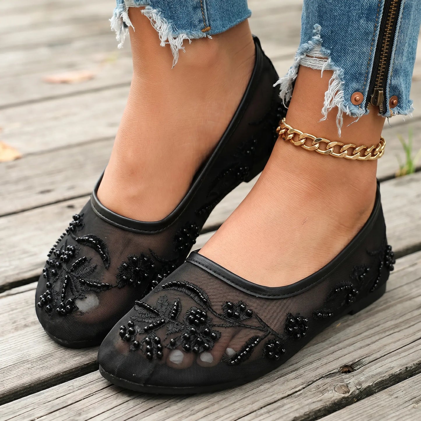 Lyraen black mesh ballet flats adorned with intricate floral embroidery, faux pearls, and matte beads, worn with distressed denim on a wooden deck.