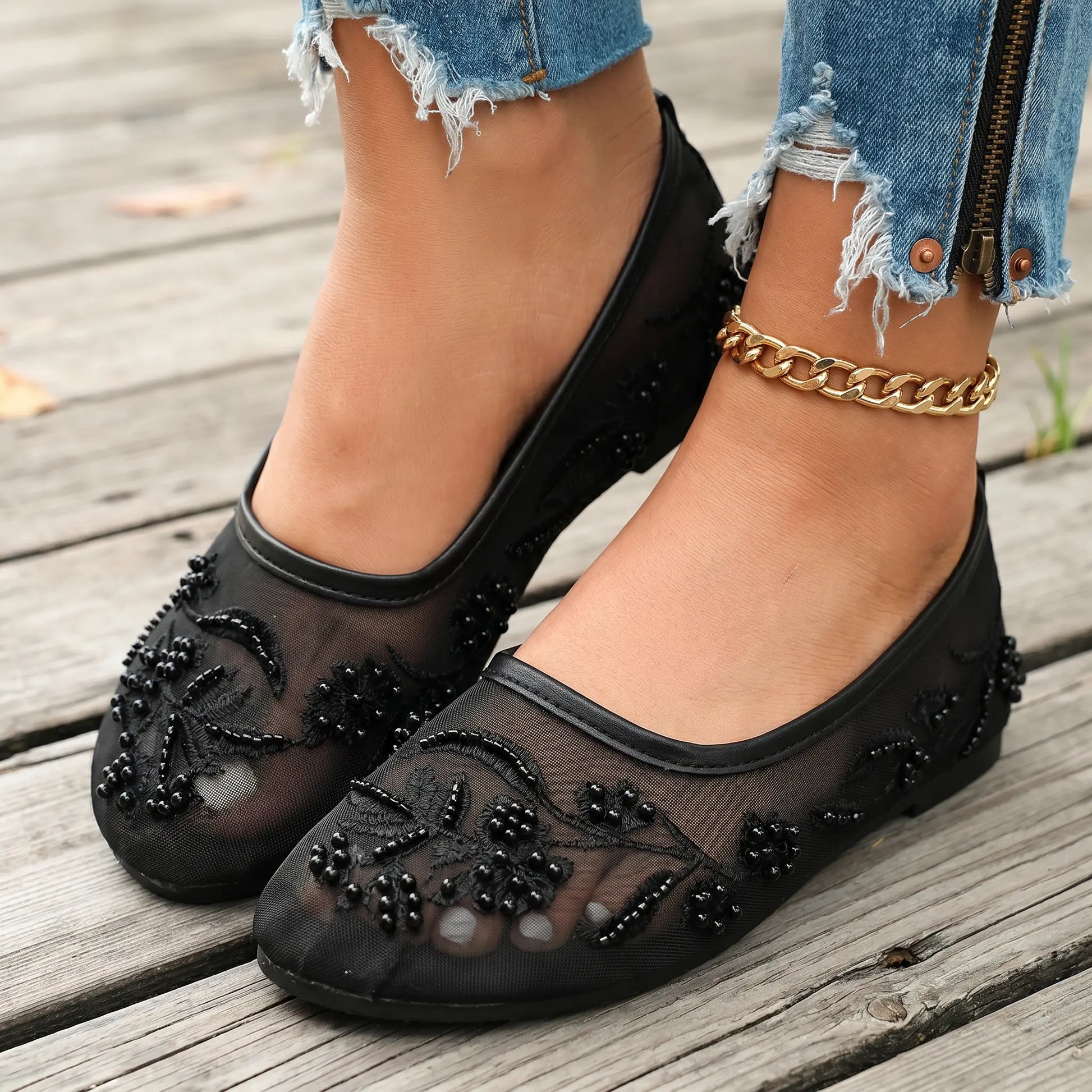 Lyraen black mesh ballet flats adorned with intricate floral embroidery, faux pearls, and matte beads, worn with distressed denim on a wooden deck.