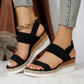 Black espadrille wedge sandals with peep-toe, textured mesh upper, elasticated straps, and a gold-tone ankle chain, worn by a model.