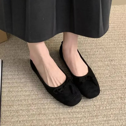 Black faux suede ballet flats, featuring a square-round toe with topstitching and a delicate knotted bow, worn on feet against a textured carpet.