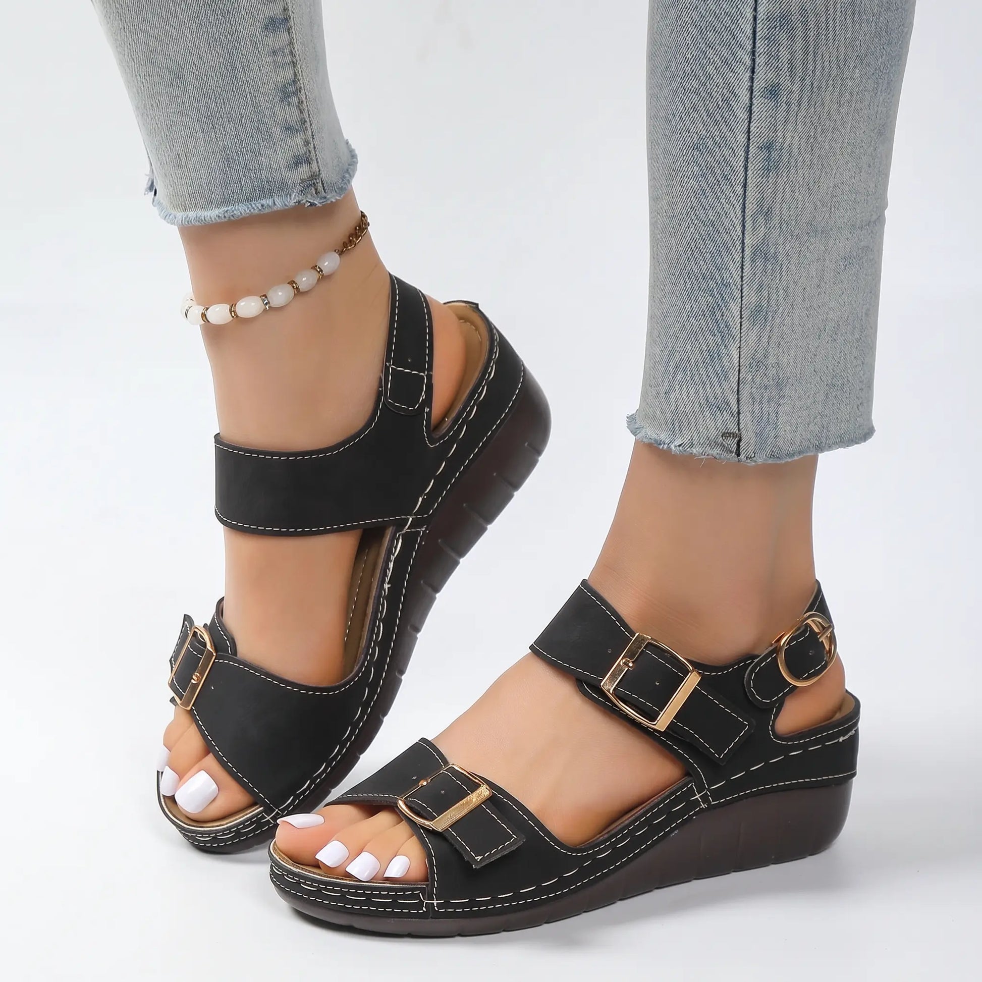Black faux suede wedge sandals for women, featuring adjustable ankle straps, gold buckles, and white contrast stitching, shown on feet in a studio setting.