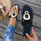 Black flat thong sandals featuring interlocking polished gold-tone rings, shown worn on a foot with distressed denim jeans and held in hand, set against a wooden deck background with floral accents.