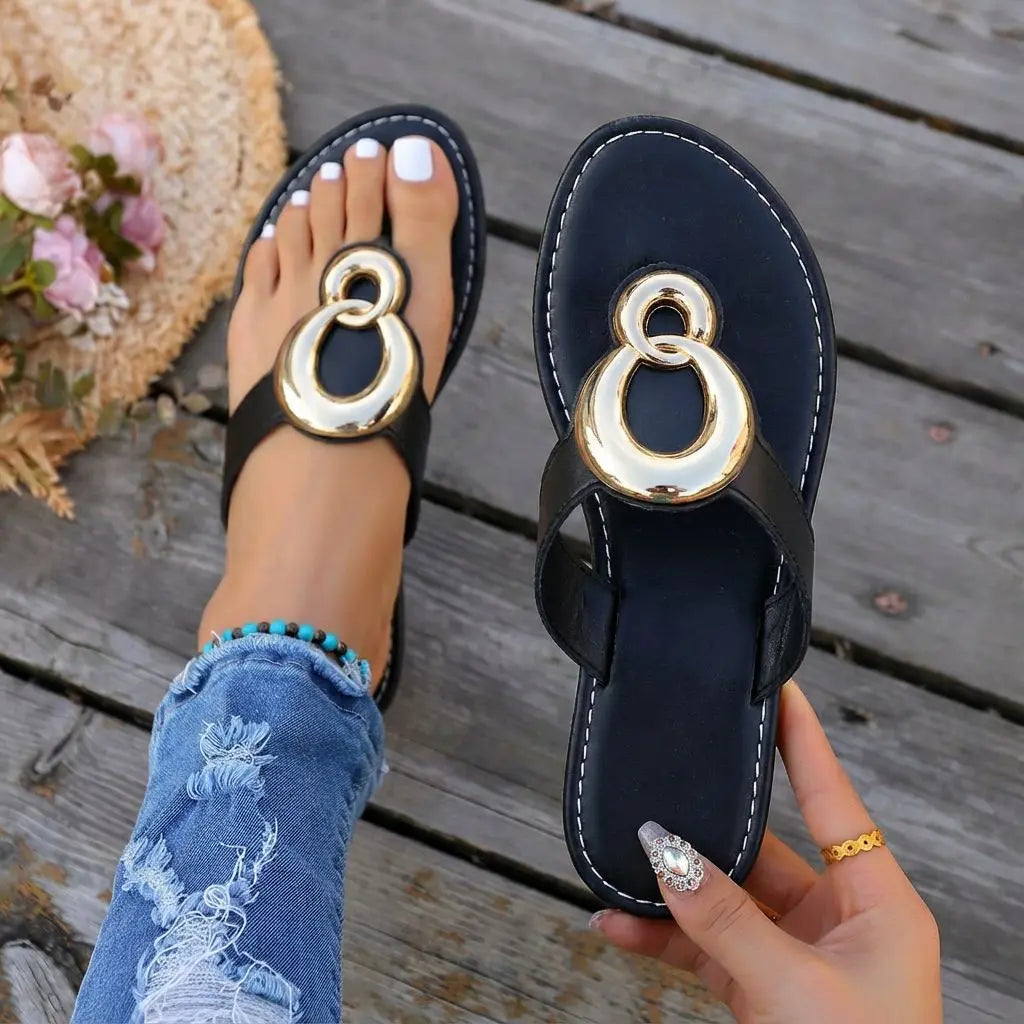 Black flat thong sandals featuring interlocking polished gold-tone rings, shown worn on a foot with distressed denim jeans and held in hand, set against a wooden deck background with floral accents.
