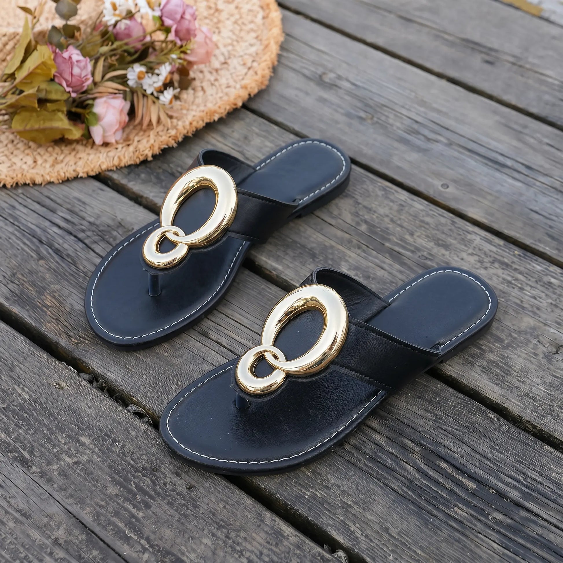 Black flat thong sandals with oversized interlocking gold-tone ring hardware and white contrast stitching, styled on a wooden deck with a straw hat and flowers.