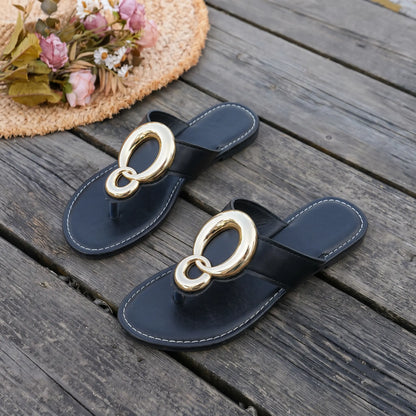 Black flat thong sandals with oversized interlocking gold-tone ring hardware and white contrast stitching, styled on a wooden deck with a straw hat and flowers.