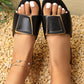 Model wearing Zovial black flat slide sandals with a prominent square gold-tone buckle detail and white pedicure, set against a natural pebble and foliage background.