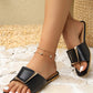 Model wearing black Zovial flat slide sandals with a distinctive square gold-tone buckle featuring a black inlay, showcased in a lifestyle setting with textured flooring.