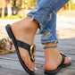 Black flat thong sandals with gold interlocking rings and white contrast stitching, worn with distressed jeans and a turquoise anklet on a wooden deck.
