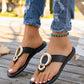 Black flat thong sandals with gold interlocking ring hardware, worn with distressed denim jeans on a wooden deck.
