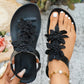 Close-up of black Dyraen bohemian flat thong sandals adorned with layered fabric floral embellishments, featuring an elasticated slingback strap and shown on a foot with white toenails and a pearl anklet.