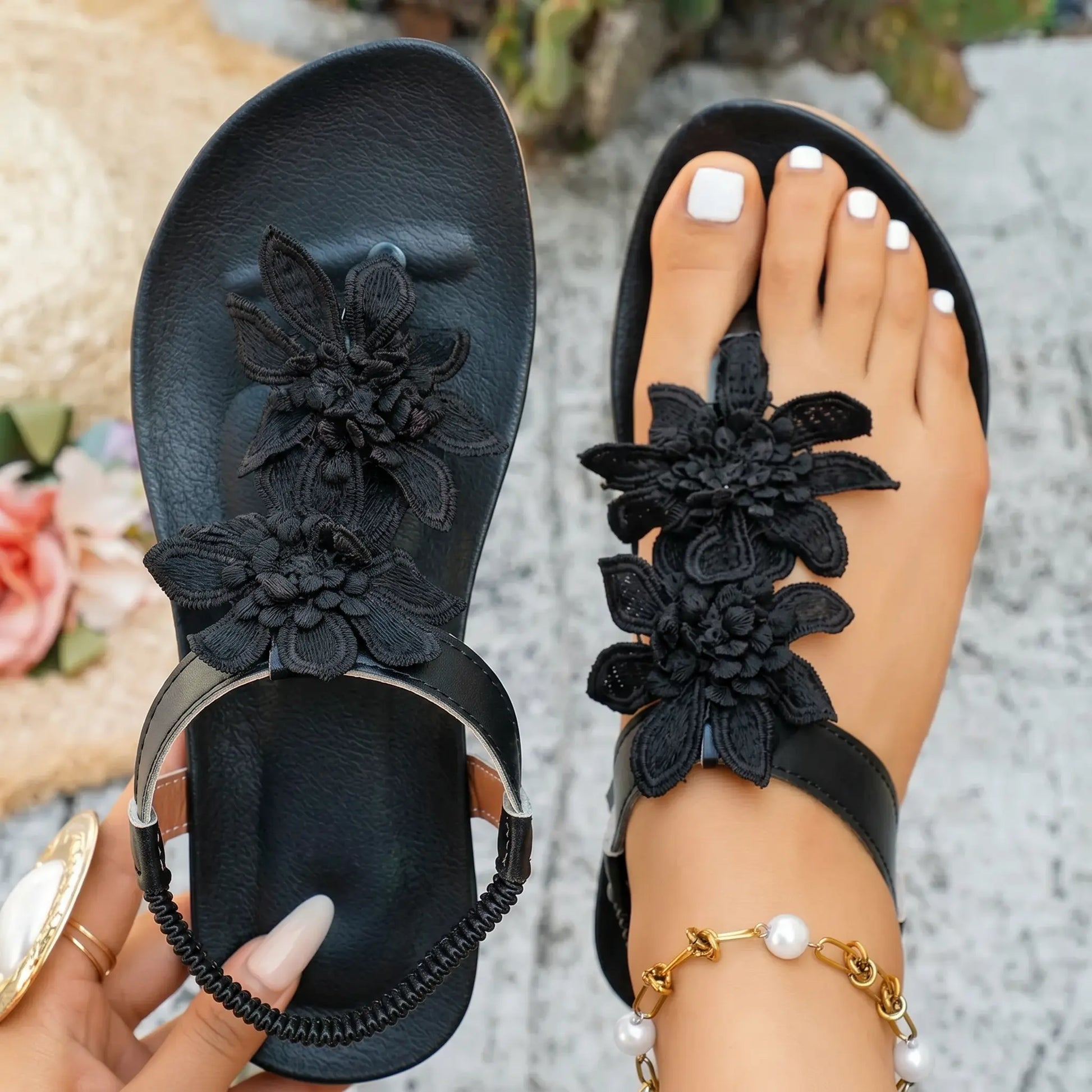 Close-up of black Dyraen bohemian flat thong sandals adorned with layered fabric floral embellishments, featuring an elasticated slingback strap and shown on a foot with white toenails and a pearl anklet.