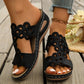 Close-up view of black bohemian wedge sandals with intricate floral laser-cut detailing on a single wide strap, worn on feet with light blue jeans and an anklet on a rustic wooden surface.