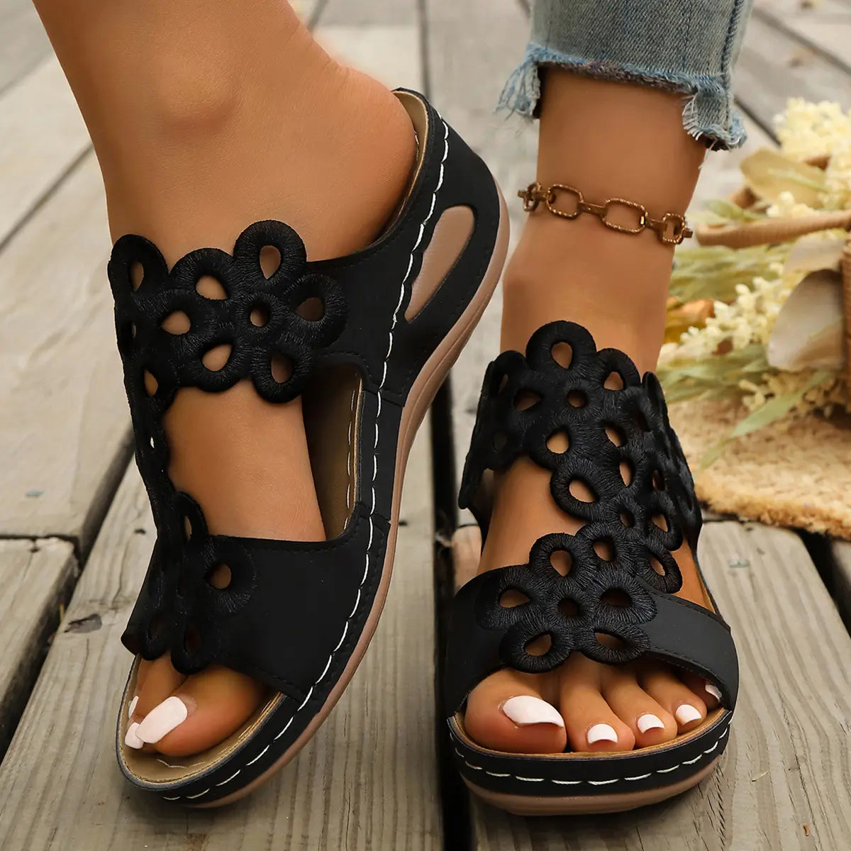 Close-up view of black bohemian wedge sandals with intricate floral laser-cut detailing on a single wide strap, worn on feet with light blue jeans and an anklet on a rustic wooden surface.