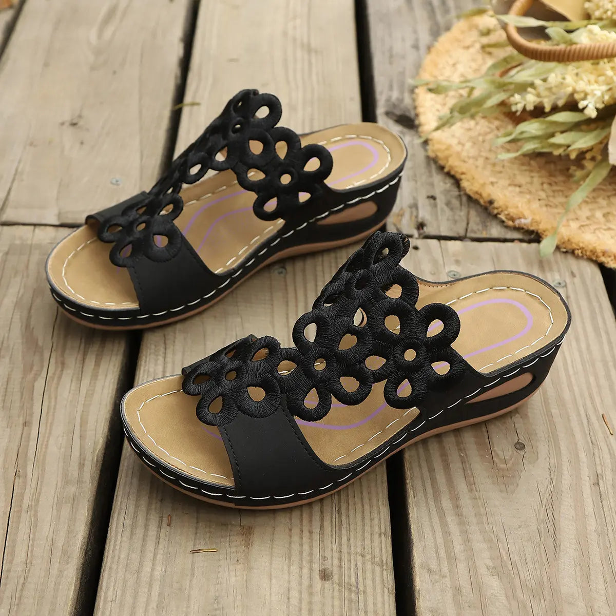 Black bohemian wedge sandals featuring intricate laser-cut floral patterns on a wide strap, displayed on a rustic wooden surface with a straw hat and dried flowers.