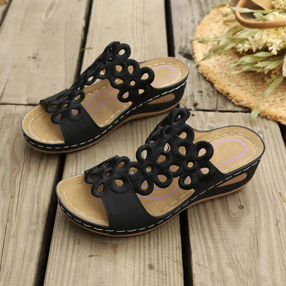 Black bohemian wedge sandals featuring intricate laser-cut floral patterns on a wide strap, displayed on a rustic wooden surface with a straw hat and dried flowers.