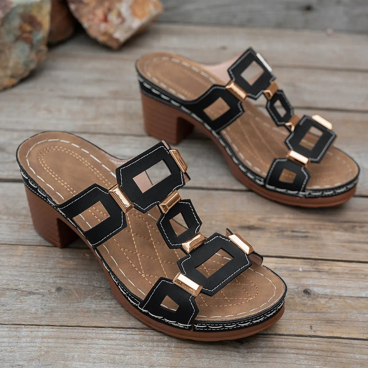 Black heeled platform slide sandals with geometric gold-tone hardware and white contrast stitching, shown from a three-quarter angle on a wooden surface.