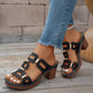 Women's black heeled slide sandals featuring a geometric strap with gold-tone connectors, worn with blue jeans on a wooden deck.