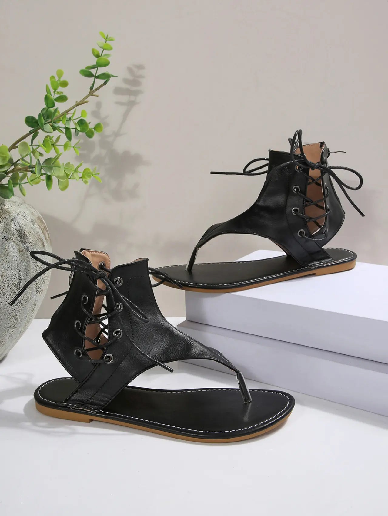 Black PU leather flat gladiator sandals with thong toe, lace-up ankle straps, and white contrast stitching around the sole.