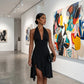 Mokazi black halter midi dress with a plunging V-neck and asymmetrical ruffled high-slit hem, worn by an elegant model in a modern art gallery.