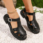 A woman's feet wearing black PU leather fisherman sandals with a jeweled buckle and geometric cut-outs, placed on white pebbles with blurred green foliage.