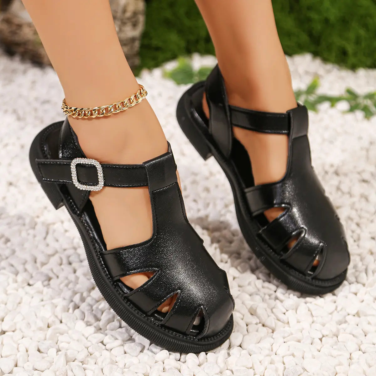A woman's feet wearing black PU leather fisherman sandals with a jeweled buckle and geometric cut-outs, placed on white pebbles with blurred green foliage.