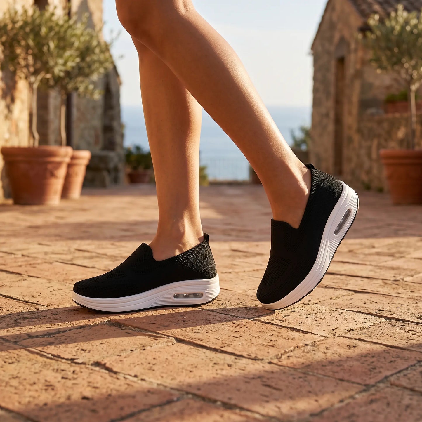 Black knit slip-on sneakers with a thick white platform and visible air cushion, worn by a woman walking on a sunlit terracotta path in a Mediterranean village.