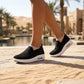 Black knit slip-on platform sneakers with visible air cushion, worn by a person walking on a textured path in a sunny desert resort.