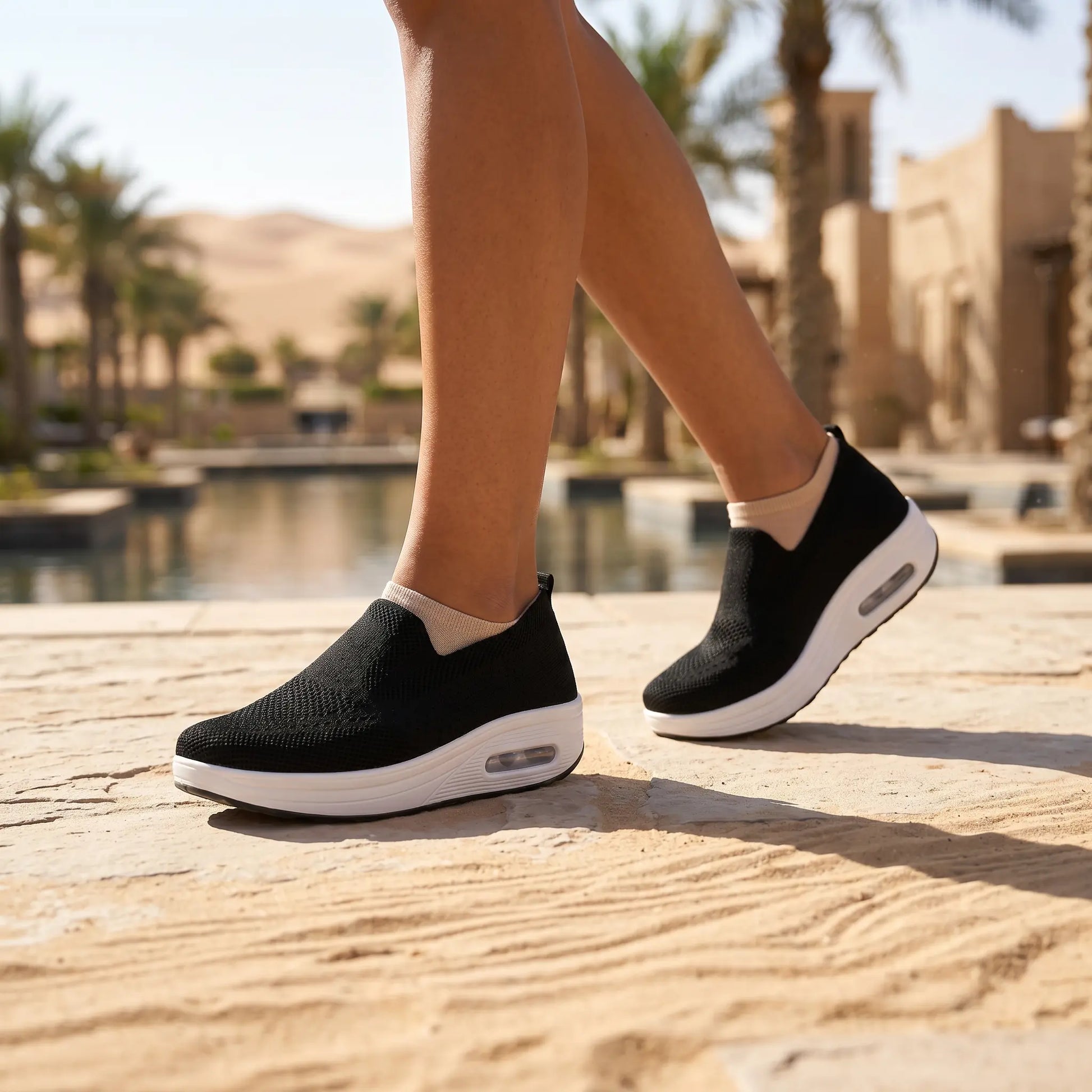 Black knit slip-on platform sneakers with visible air cushion, worn by a person walking on a textured path in a sunny desert resort.