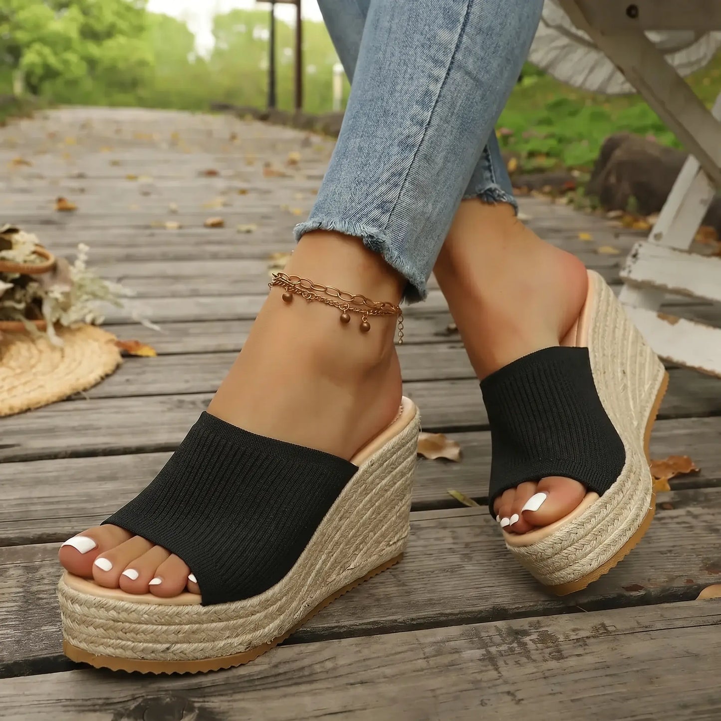 Black knitted espadrille wedge sandals with open toe and jute-wrapped platform worn by a woman on a wooden boardwalk, styled with blue denim jeans.