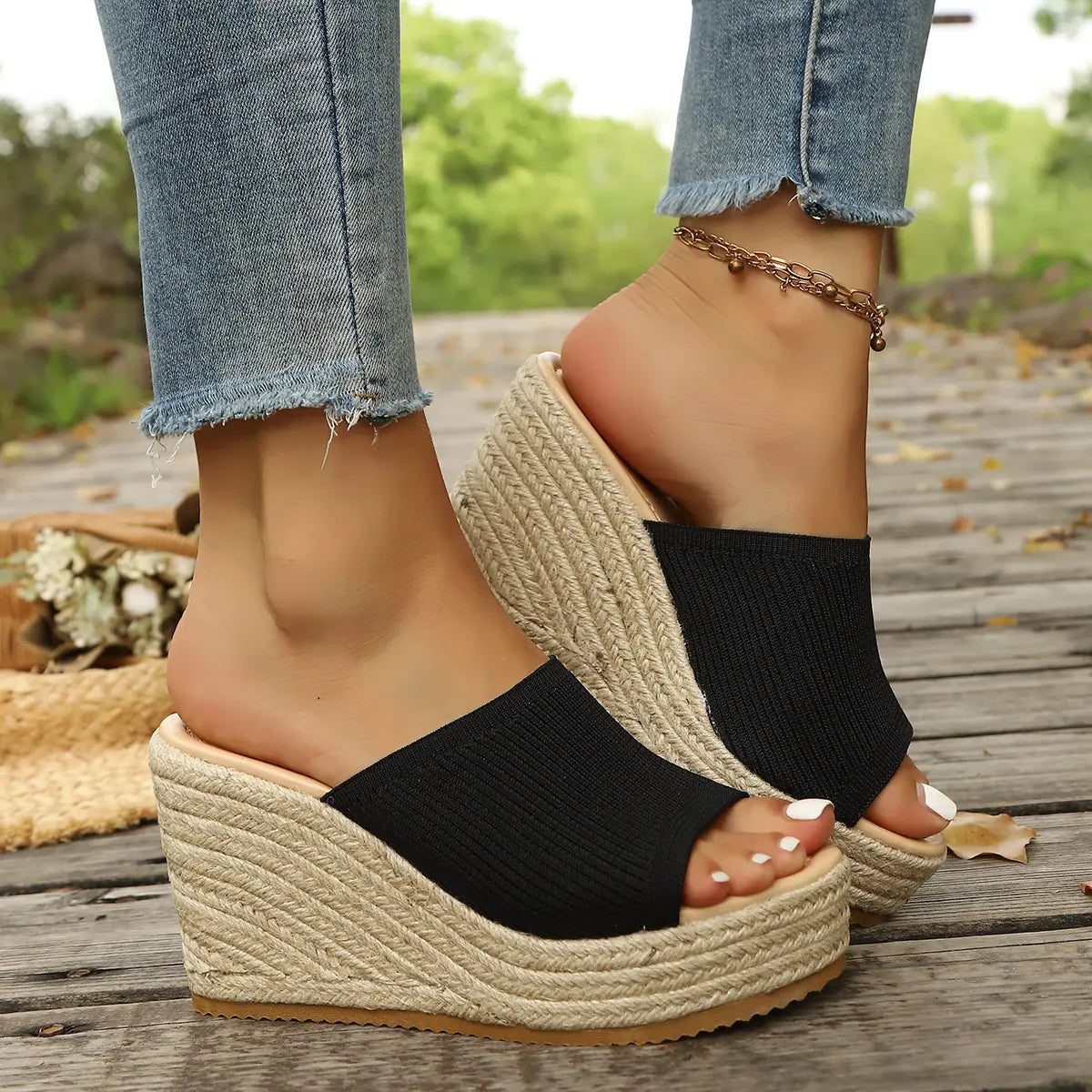 Black knitted espadrille wedge sandals with an open toe and substantial platform, shown on a woman's feet on a rustic wooden boardwalk, complemented by blue denim jeans and natural surroundings.