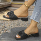 Black low heel slip-on slide sandals featuring a prominent twisted knot detail on the vamp, shown on a woman's feet in an outdoor setting with a blurred background of greenery and urban elements.