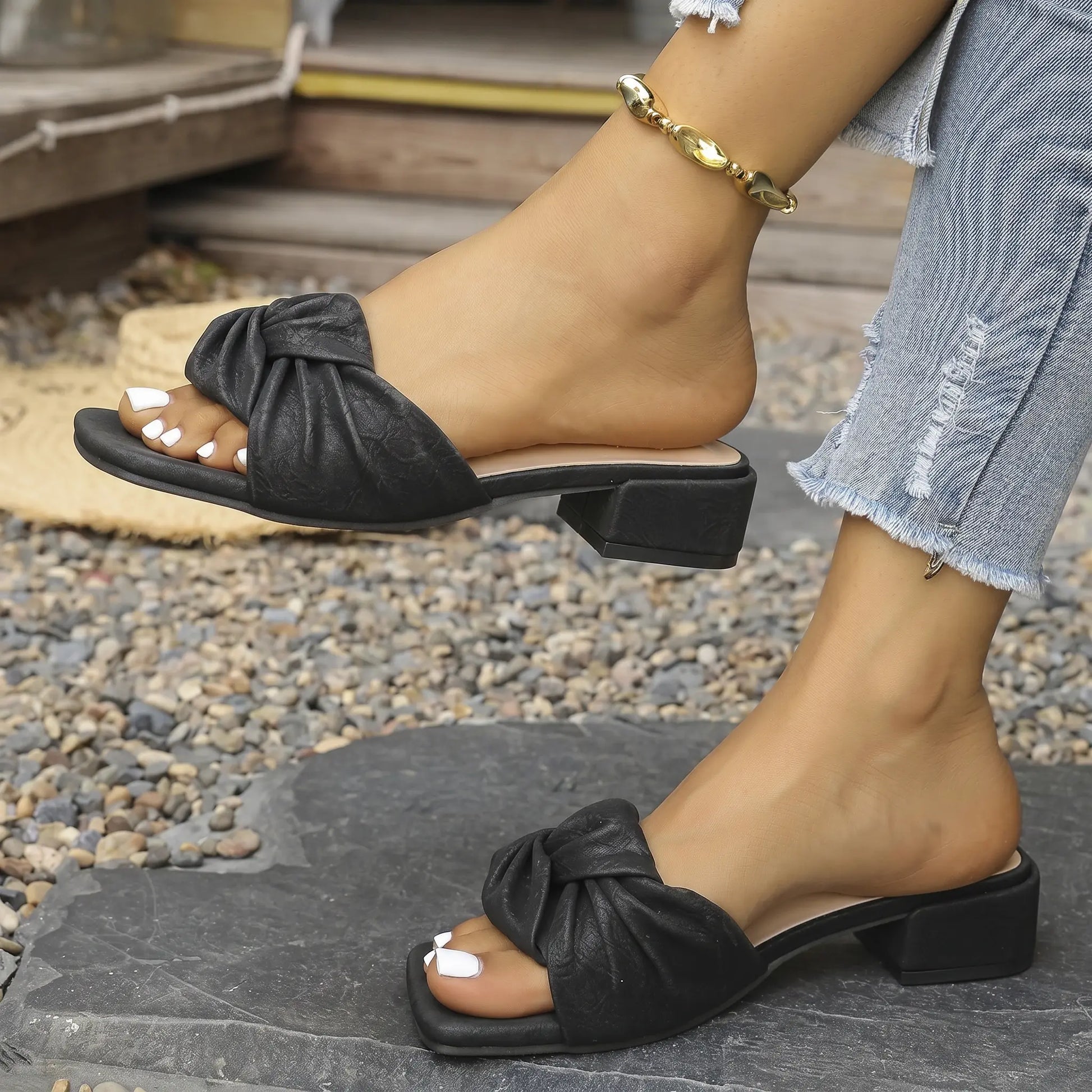 Black low heel slip-on slide sandals featuring a prominent twisted knot detail on the vamp, shown on a woman's feet in an outdoor setting with a blurred background of greenery and urban elements.