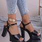 Black Kylos platform high heel sandals with an ankle strap and sparkling charm are worn by a model with distressed denim jeans on a reflective surface with disco ball elements.