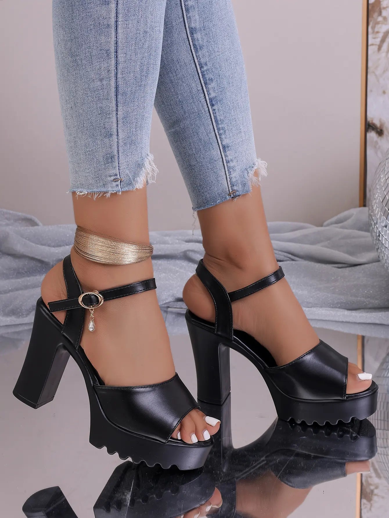 Black Kylos platform high heel sandals with an ankle strap and sparkling charm are worn by a model with distressed denim jeans on a reflective surface with disco ball elements.