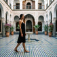 A woman gracefully walks through an ornate courtyard, wearing a black Kynso Premium Modal Slip Dress. The dress features a scoop neckline, delicate spaghetti straps, and a flowing A-line silhouette.