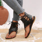 Black faux leather gladiator sandals with a thong toe post and lace-up ankle straps, worn by a woman in grey jeans against a textured tile and stone background.