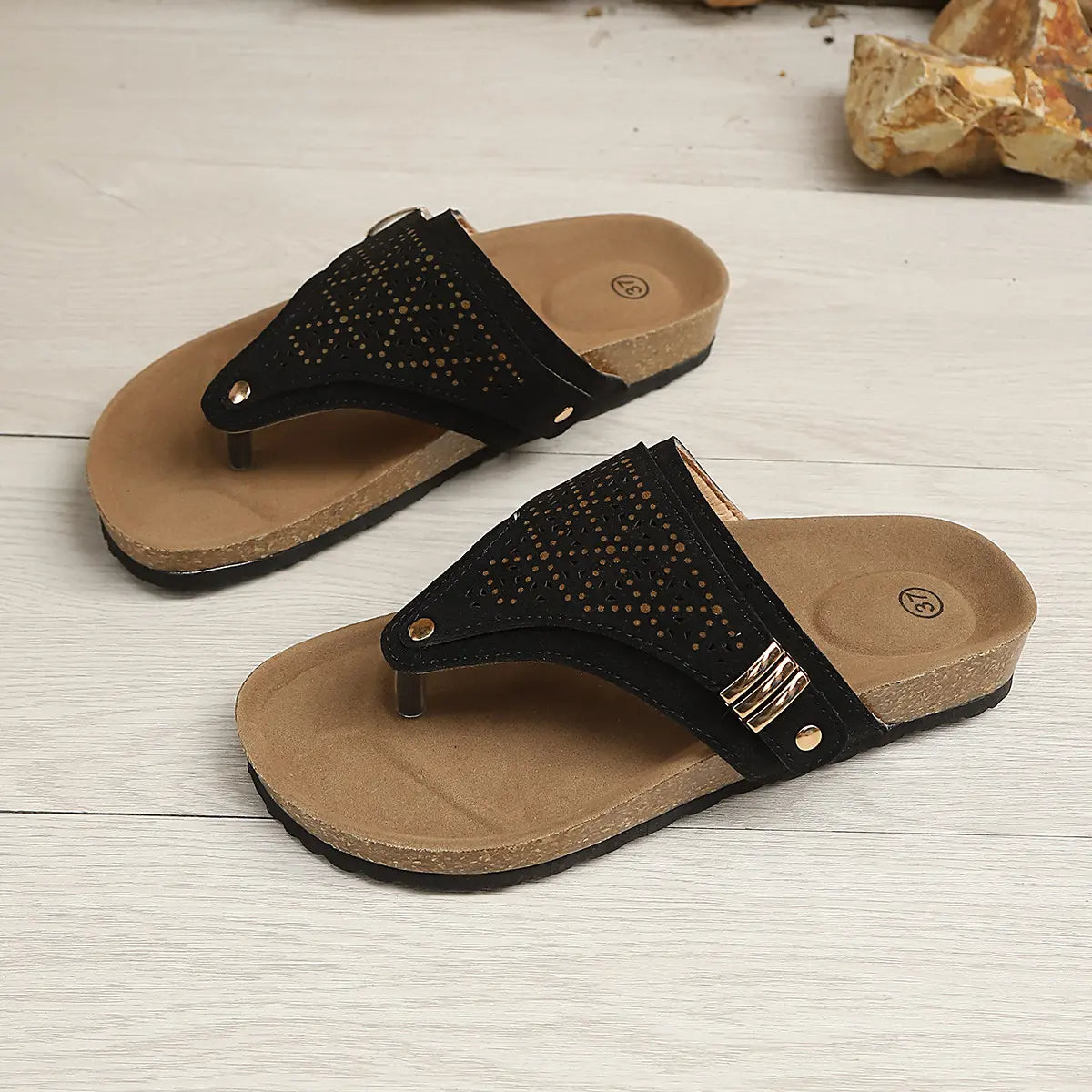 Black suede-like thong platform sandals with intricate laser-cut detailing, a cork-patterned footbed, and gold-tone buckle accents, presented at a three-quarter angle on a light wooden surface.