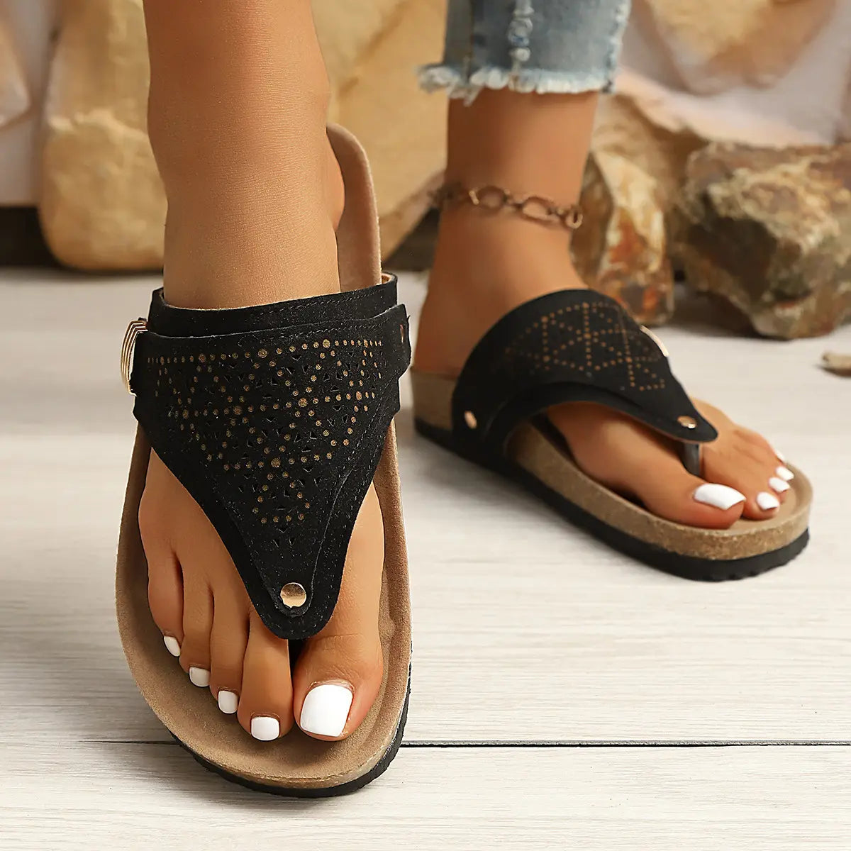 Black suede-like PU thong sandals with intricate laser-cut patterns and a cork-textured footbed, worn on feet with white toenails, showcasing the adjustable gold buckle and platform sole against a naturalistic background. Elegant summer footwear.