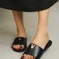 Woman wearing black smooth polyurethane leather slide sandals with a polished gold-tone metal V logo embellishment, shown from a top-down perspective on a neutral tiled floor.