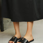 Black smooth polyurethane leather slide sandals for women, featuring a gold 'V' logo emblem on the strap, worn with a black skirt.